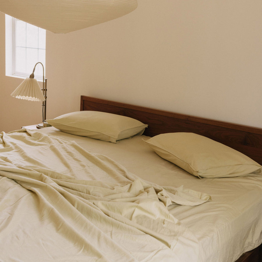 A bright bedroom made with Límon / light yellow sheets on a dark wooden bed and a lamp in the background