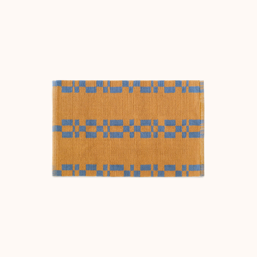 A rectangular, hand looped bathmat with a geometric pattern featuring blue and camel colors.