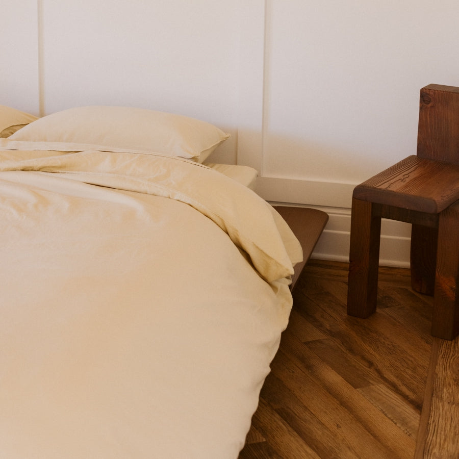 A close-up of a bed with light yellow bedding and pillows, positioned next to a dark brown wooden stool on a polished wood floor against a white paneled wall.