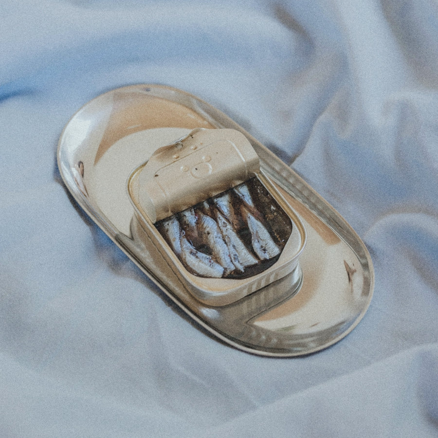 A small oval metal tin with its hinged lid open, showing several dark fish fillets inside, placed on a matching oval metal tray over light blue fabric.