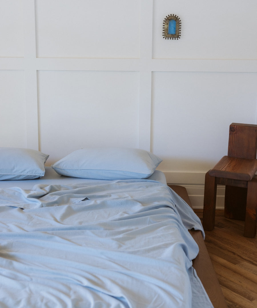 Light blue bedding, including a duvet cover and two pillows, styled on a bed in a bedroom with white paneled walls, a small blue-framed mirror, and a wooden stool.