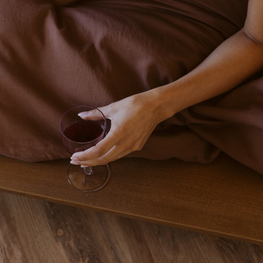 A close-up lifestyle shot of a person resting in bed, holding a stemmed glass of red wine on a wooden bedside table. The bedding is a rich, warm brown color, and the background shows a wooden floor.