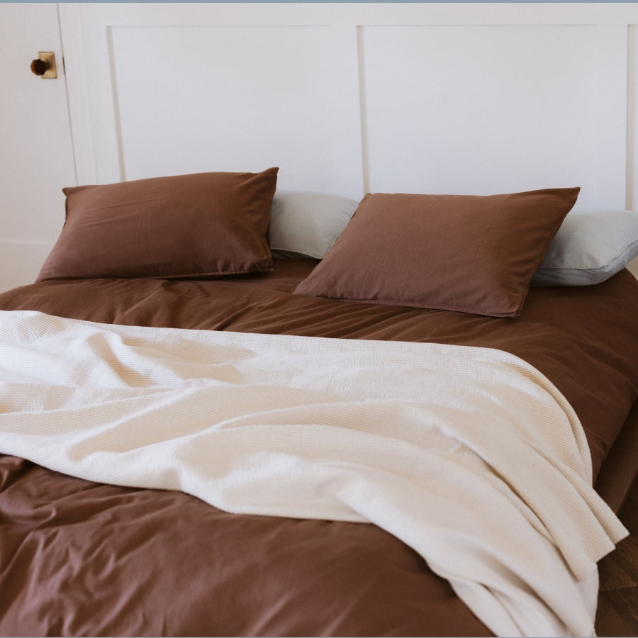 A bed dressed in a brown bedding set with two brown pillows, a cream throw blanket draped across the front, and a white paneled headboard in the background.