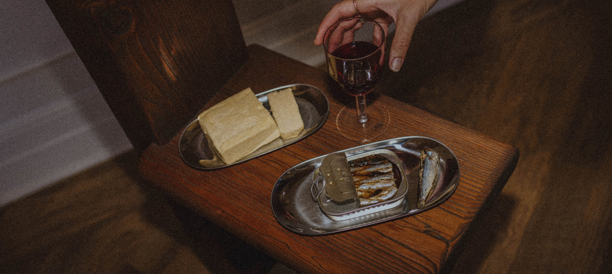 A hand holding a glass of red wine over a rustic wooden table, with two silver oval trays holding butter and tinned fish. The scene is dimly lit with a warm, cozy atmosphere.