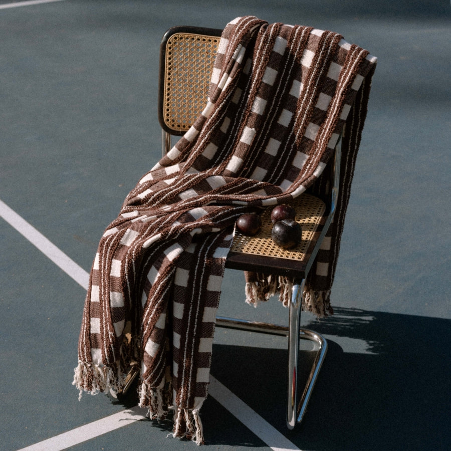 A brown and cream checkered throw blanket with fringe is draped over a modern cane-back chair on an outdoor tennis court.