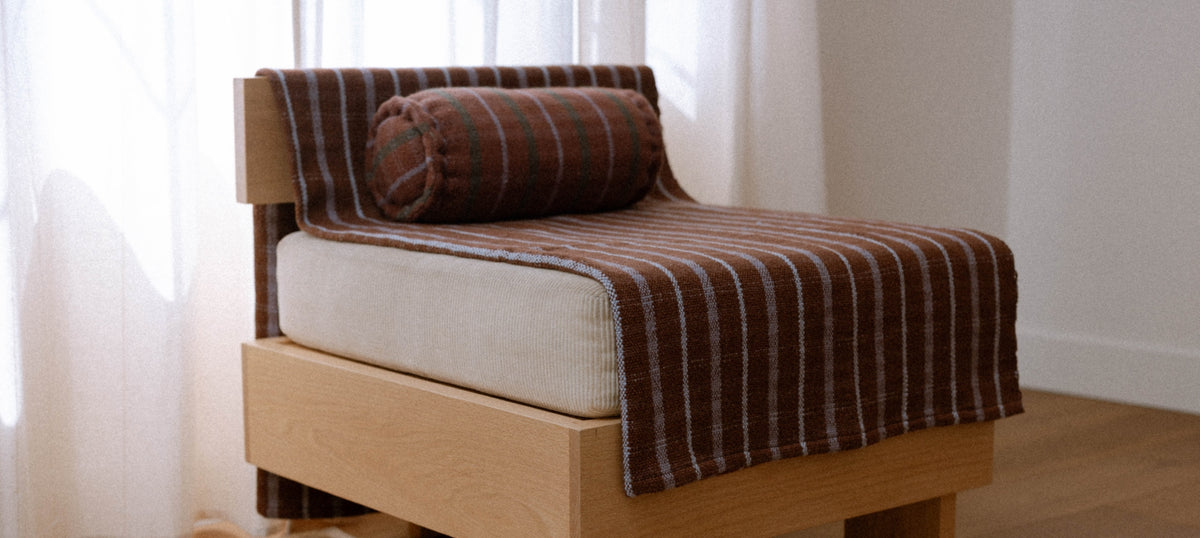 A wooden chair with a brown and purple striped blanket and matching striped cylindrical pillow, set in a bright room with sheer white curtains.