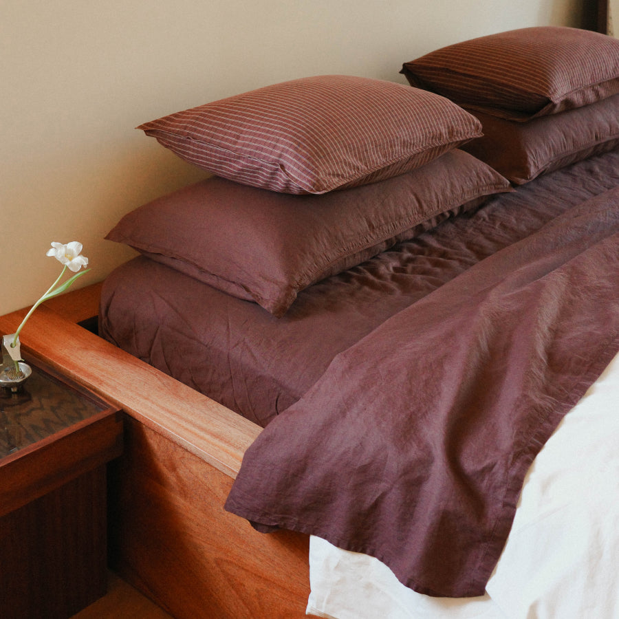 A bed dressed in coordinated brown bedding, including a duvet cover, fitted sheet, and multiple pillows, styled in a warm bedroom setting with a wooden headboard and bedside table.