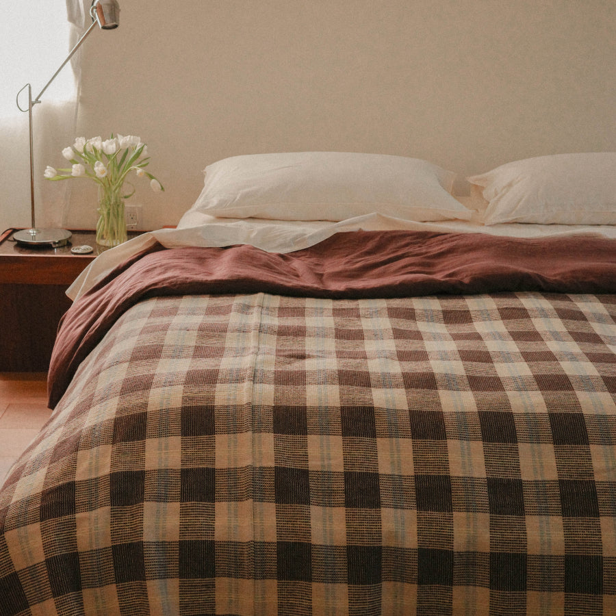A bed dressed in a brown and beige plaid checkered blanket, with a dusty rose throw folded across the top, in a softly lit bedroom setting.