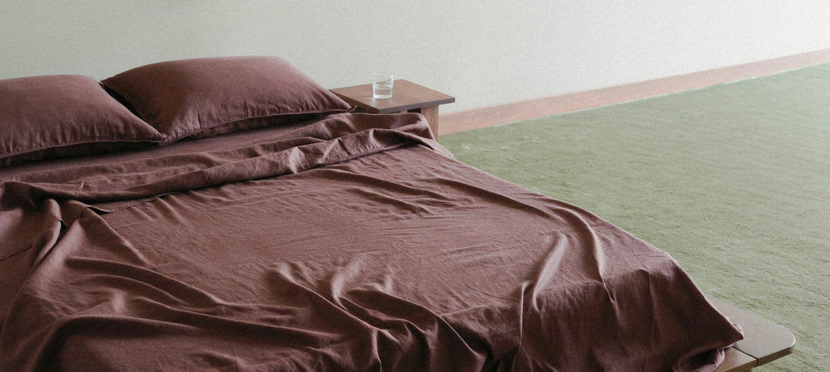 A bed dressed in dark brown bedding, including a duvet cover and two pillows, styled in a minimalist bedroom setting with a light green floor and a small wooden side table.