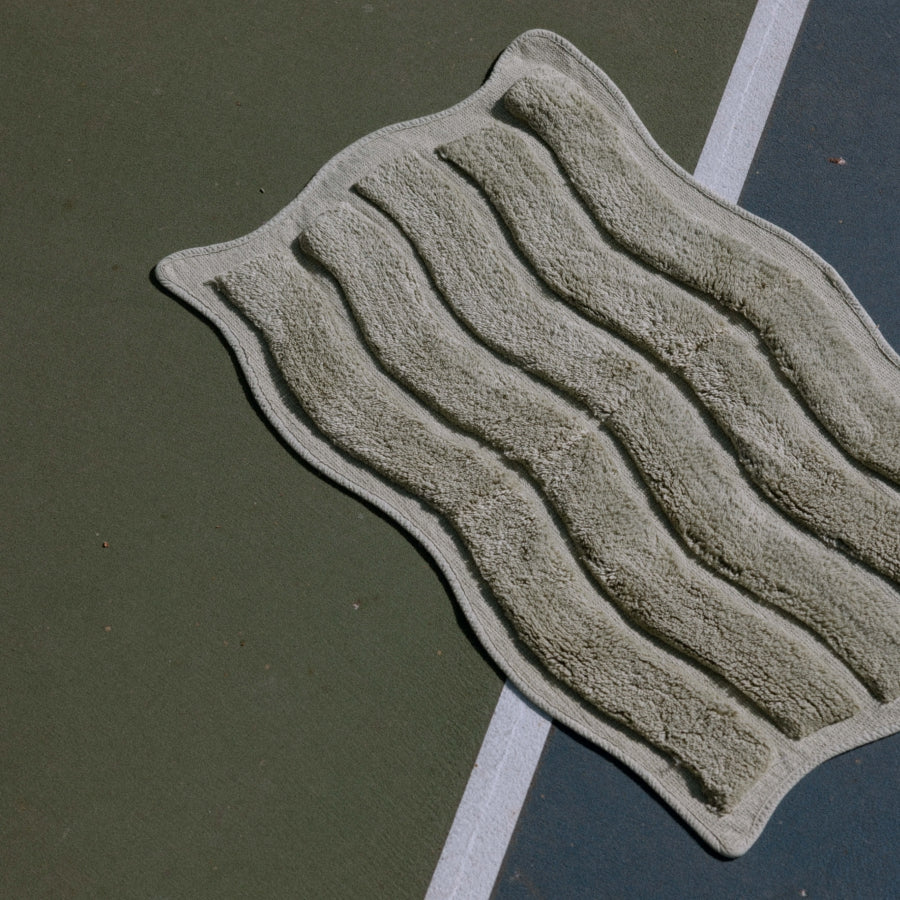 A textured, ribbed bath mat in a muted green color, shown laid out diagonally on an outdoor tennis court.