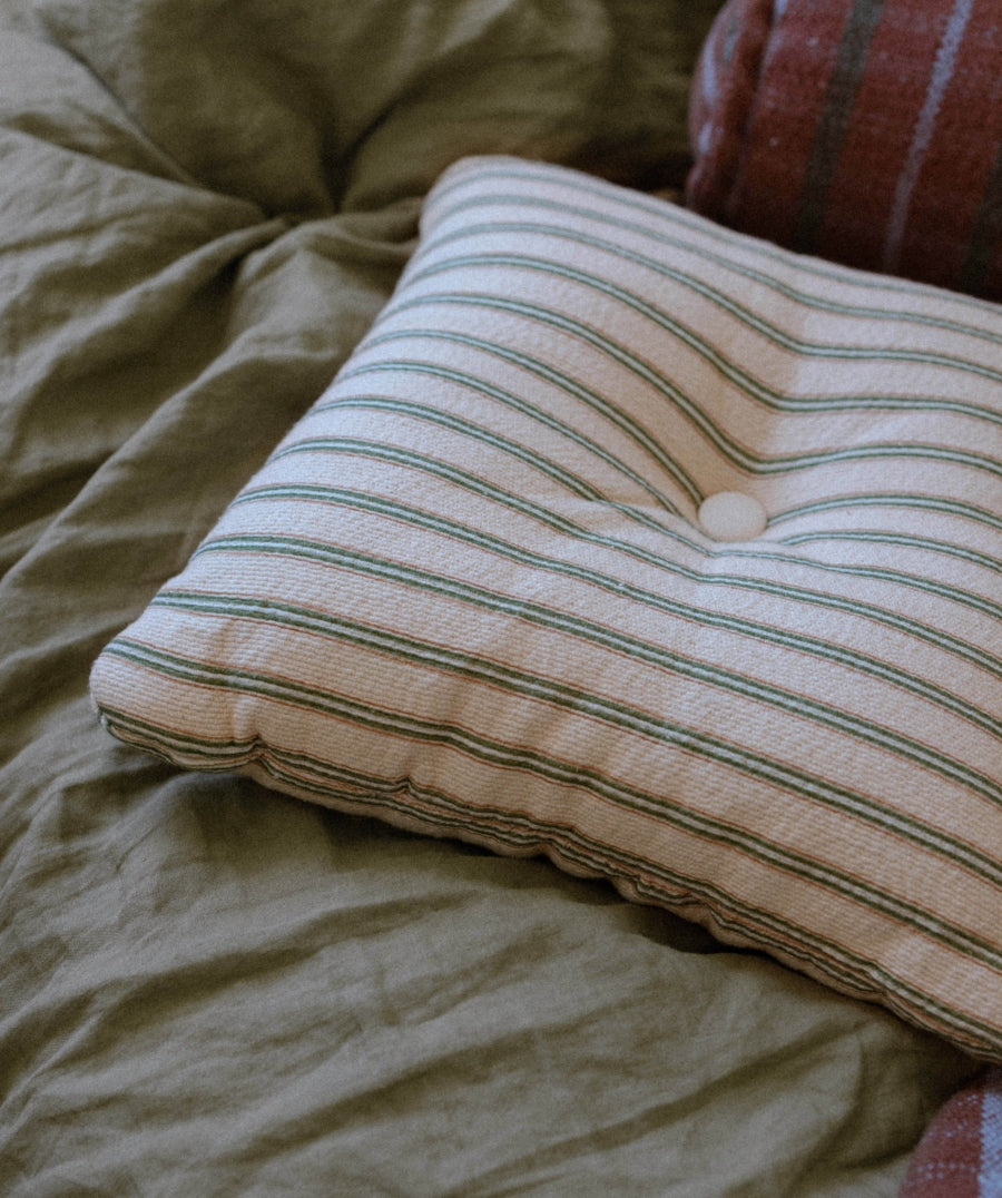 A square tufted pillow with thin green and beige stripes, resting on a textured olive green linen bedspread.