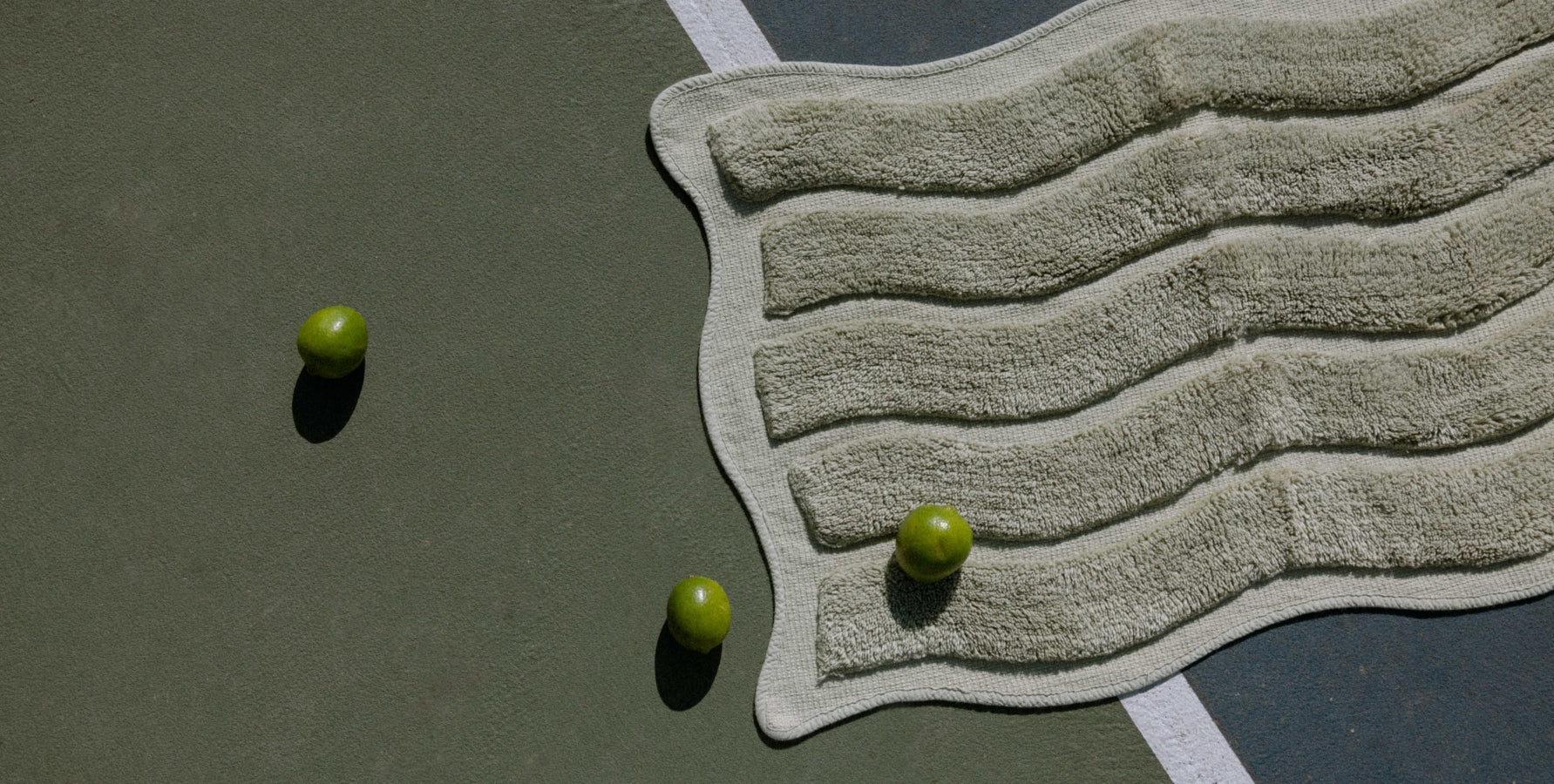 A textured wavy green bath mat, styled on a dark green tennis court with three green limes.