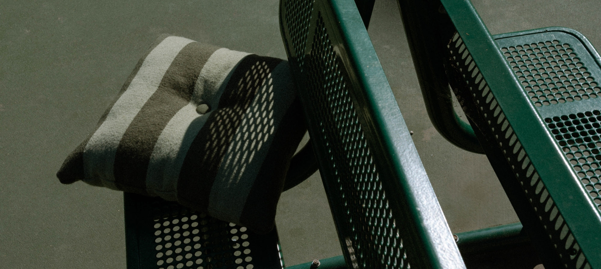 A close-up of a striped green pillow resting on a green metal mesh bench.