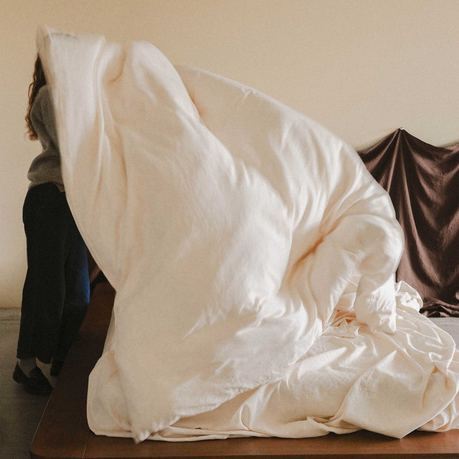 An obscured woman is shaking out a cream colored duvet cover, perhaps making the bed. 