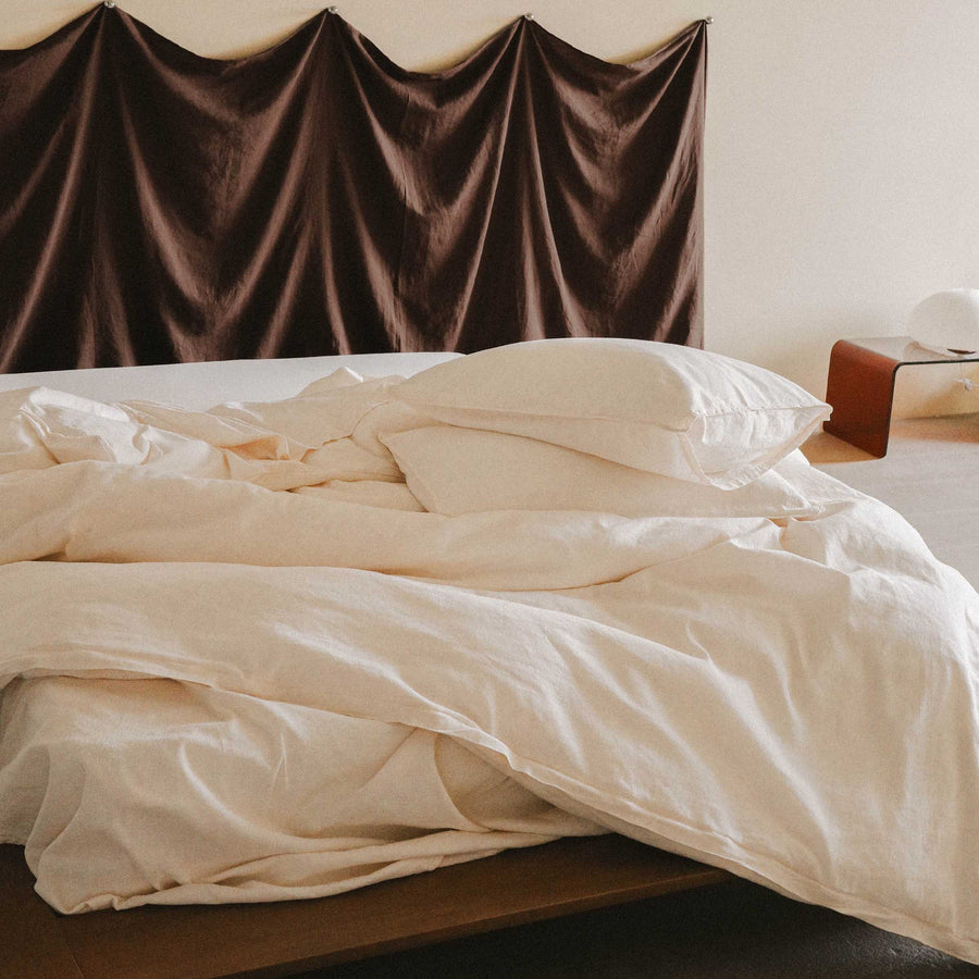 An unmade cream / white bed with two pillows and a nightstand nearby with a paper lantern 