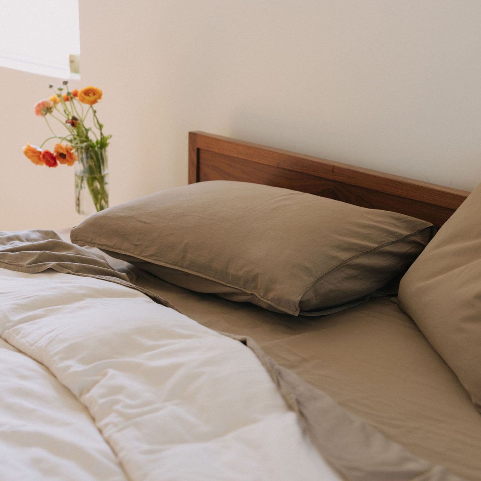 A bed dressed in beige sheets and pillows and a white duvet on a wood bed with a base of flowers in the background