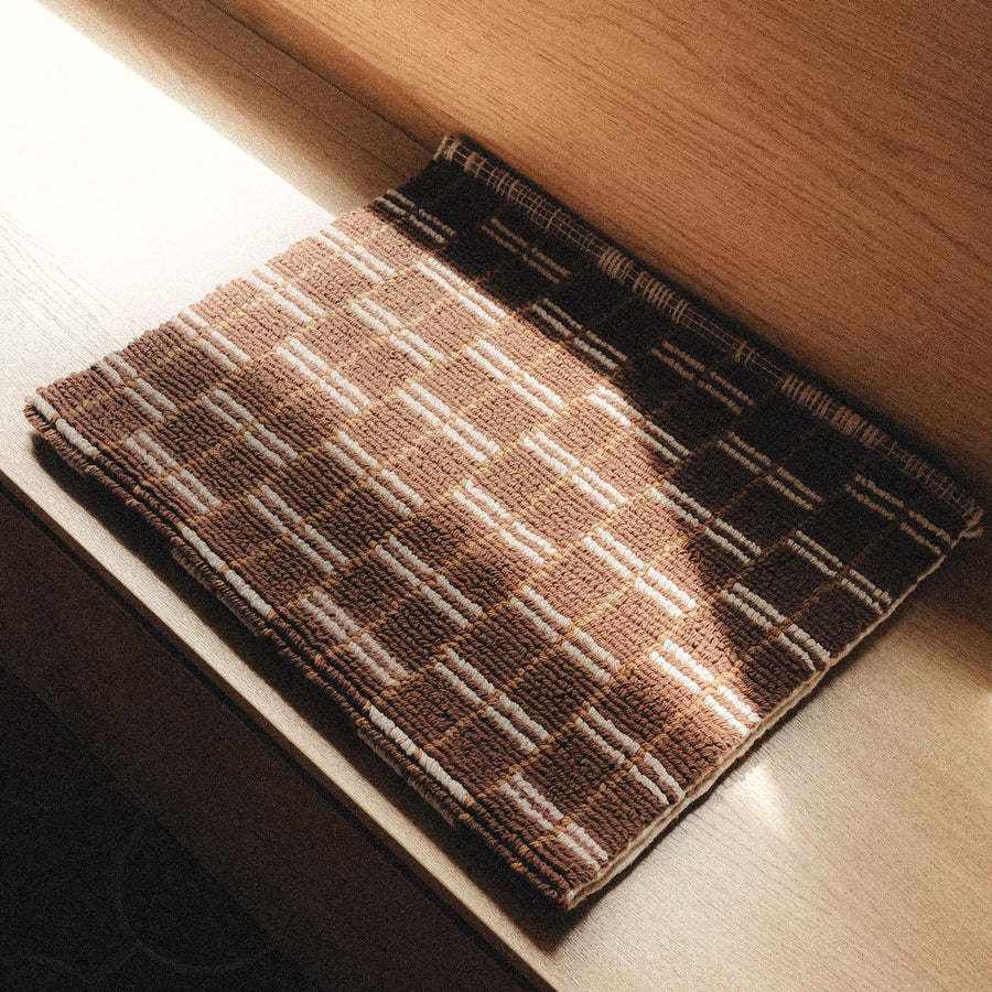 A brown bathmat with a tan geometric design folded in half and sitting on a wooden bench.