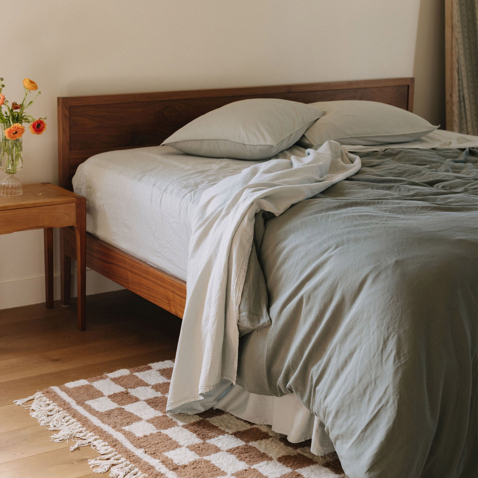 A bedroom setting with a brown and white checkered rug and a bed made with sheets of pale blue / Mist and a deeper green duvet cover