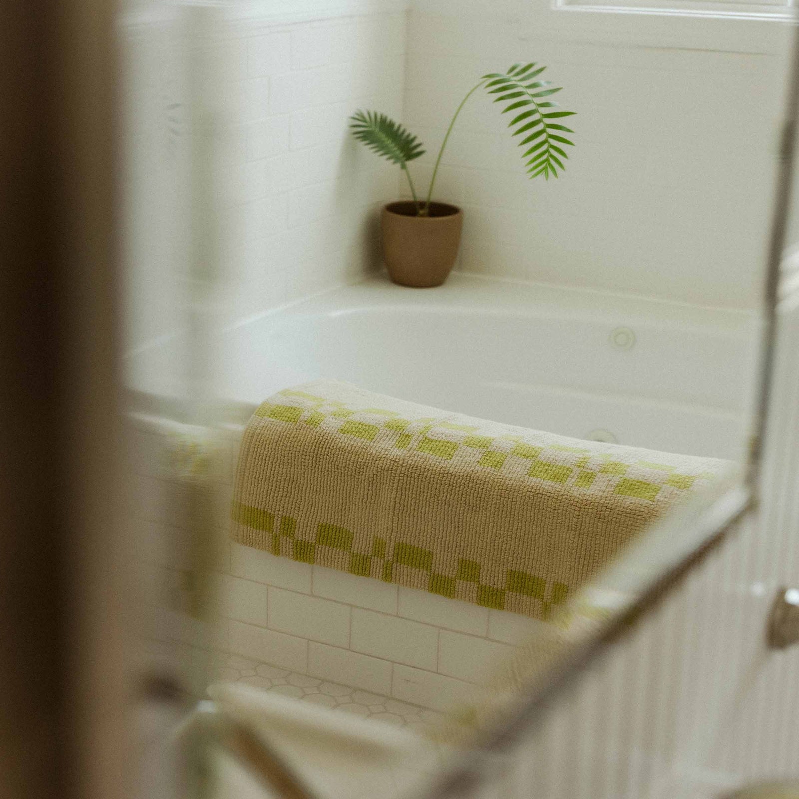 A bathmat with a lime geometric pattern and a beige background draped over a tile tub with a plant in the corner.