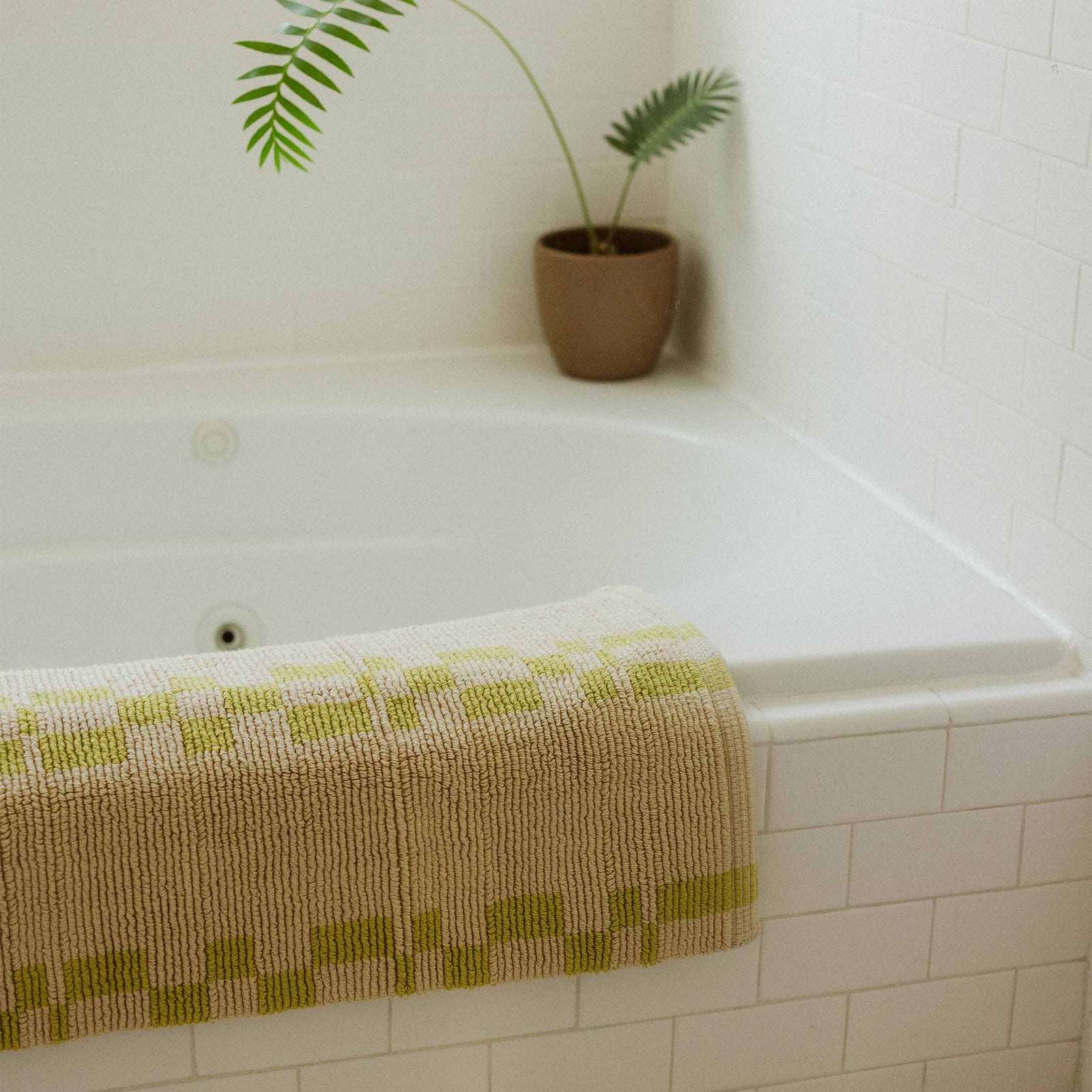 A bathmat with a lime geometric pattern and a beige background draped over a tile tub with a plant in the corner.