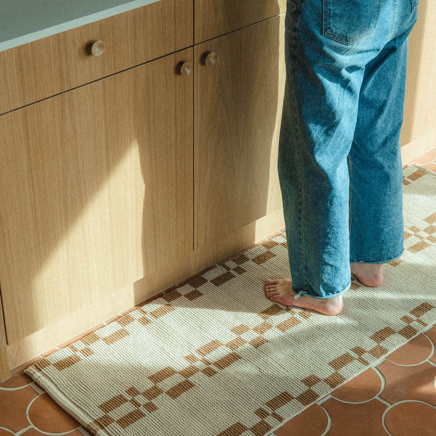 Someone wearing jeans standing on a brown and beige runner with a geometric pattern on a terracotta tile floor of a kitchen.