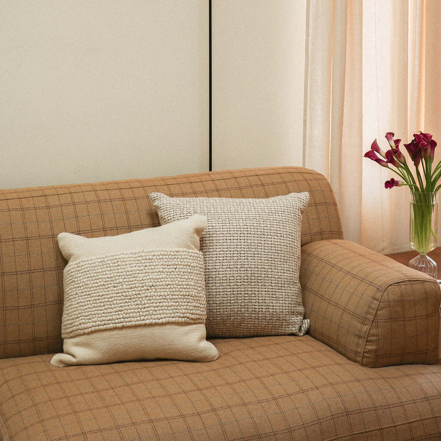 A cream textured throw pillow and a tan patterned throw pillow on couch with flowers on a side table. 
