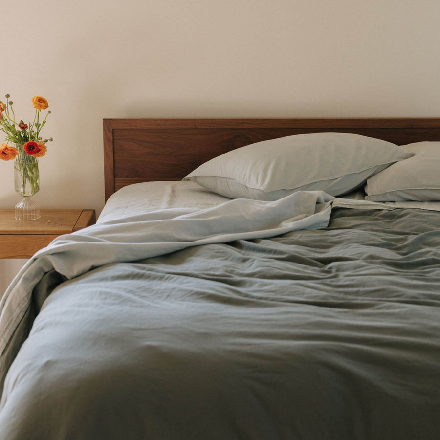 A bed with light blue sheets and a green duvet cover in a bedroom setting with a nightstand dressed with a vase and flowers 