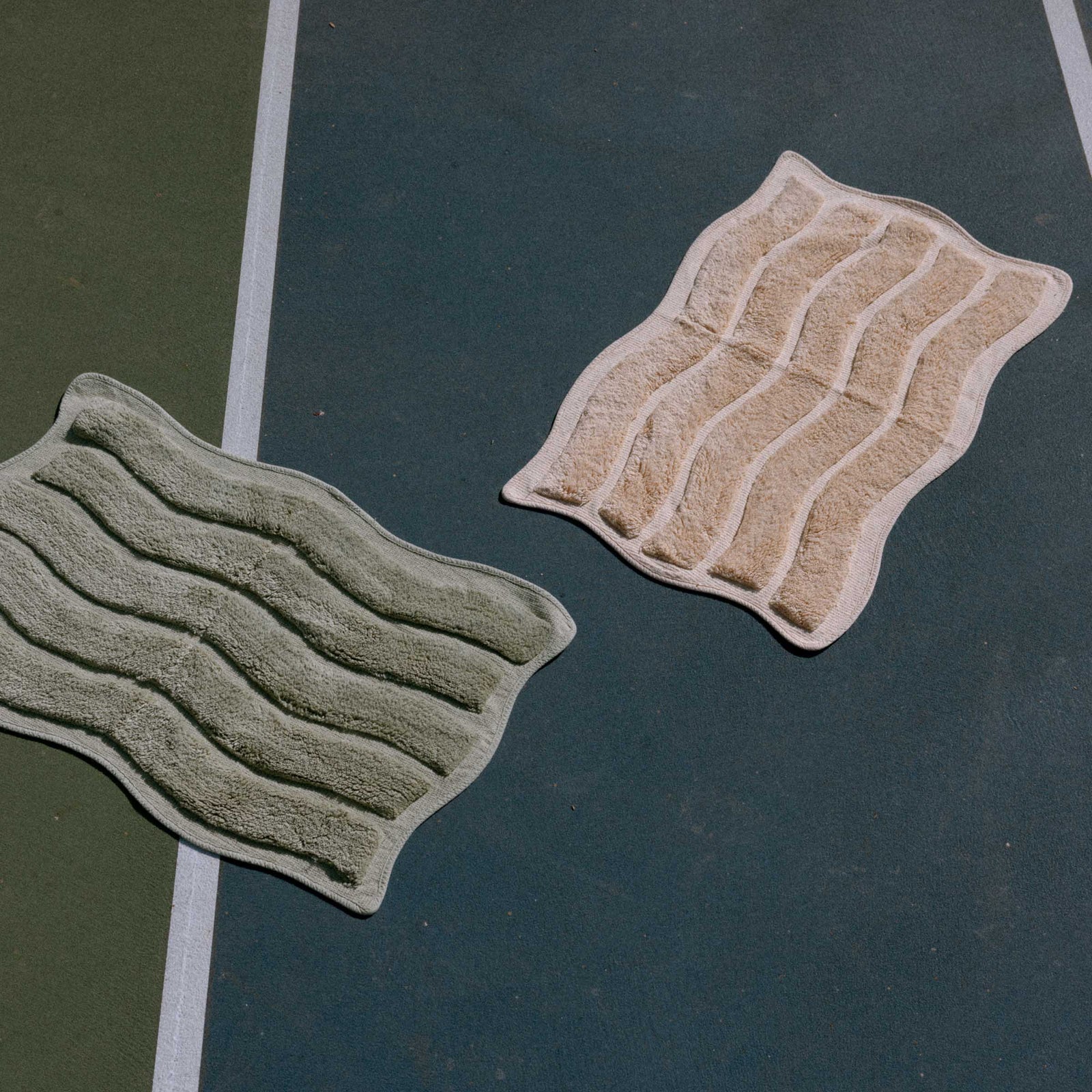Two textured bath mats, one green and one beige, on the dark surface of a tennis court.