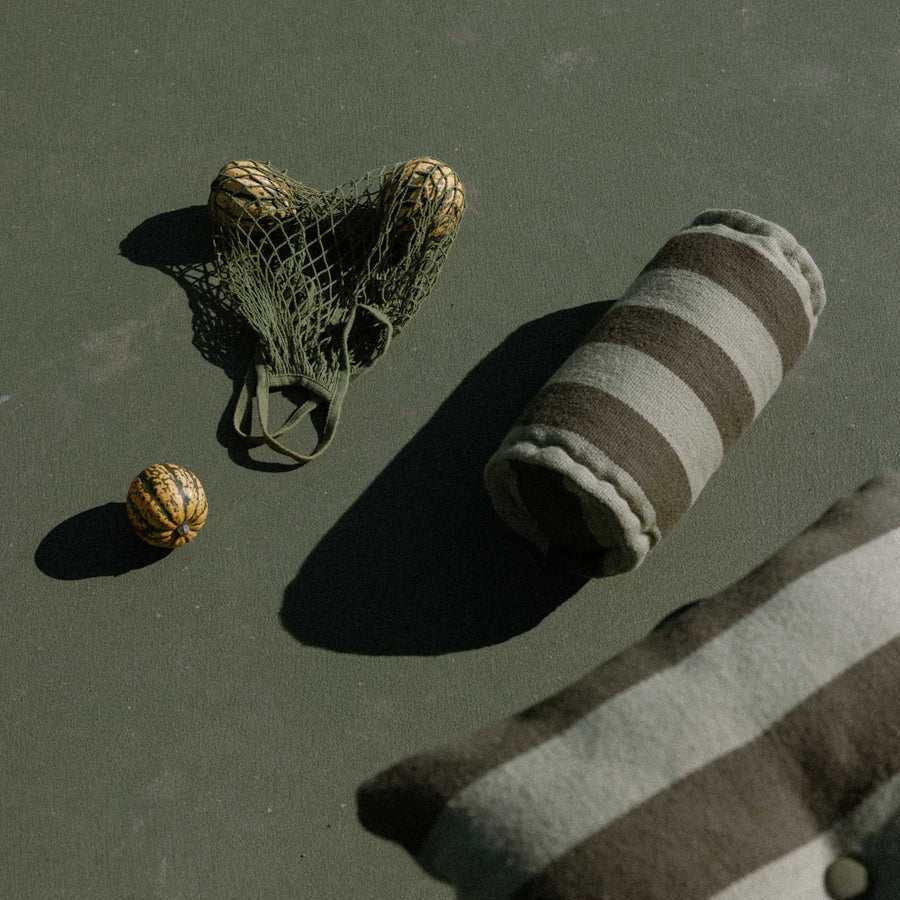 Net bag with fruit next to a striped bolster and pillow in two shades of green on a tennis court.
