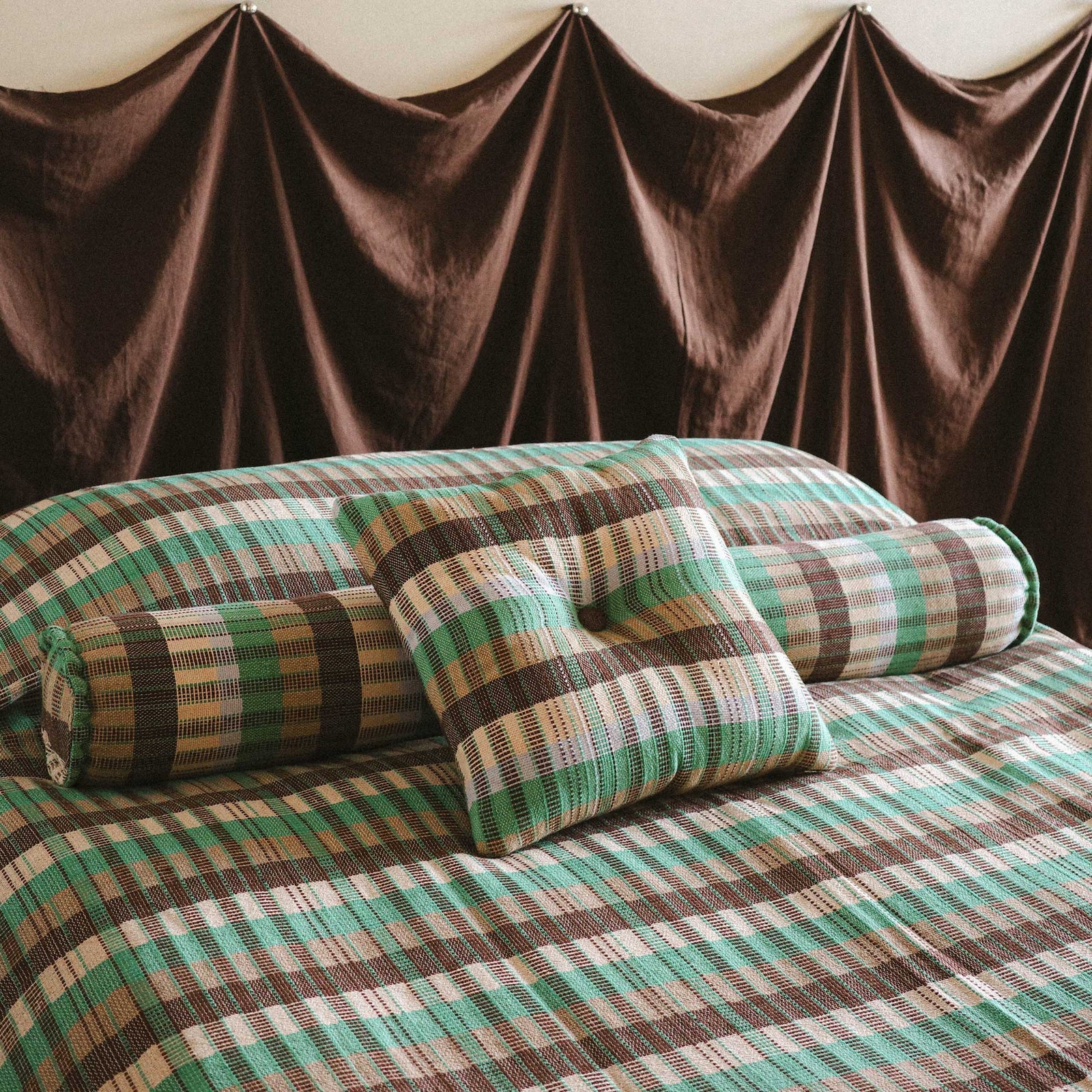 A bed styled with the Yara bed cover, Yara bolster and Yara throw pillow. There is a dark brown fabric headboard draped in the background