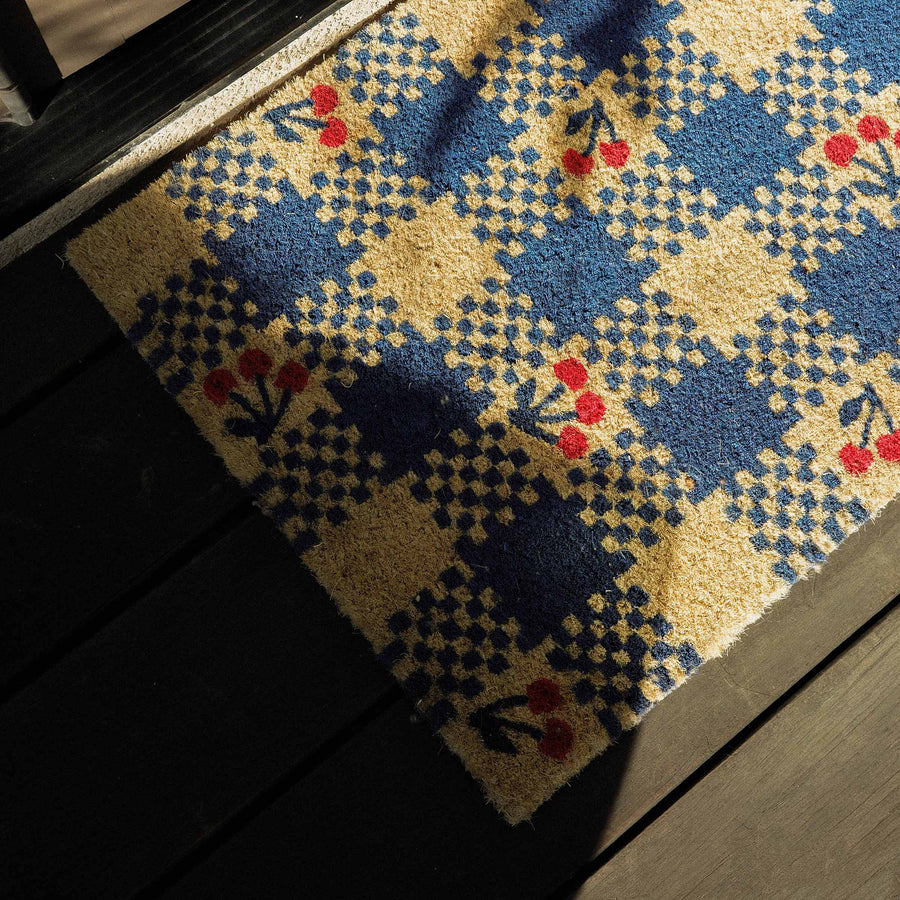 Decorative doormat with a blue checkered pattern and red cherries on a wooden floor.