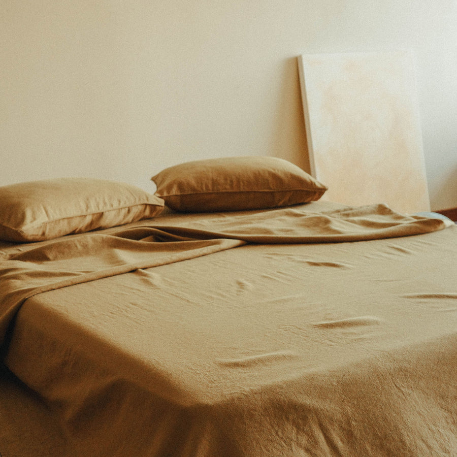 Bed with brown bedding and pillows against a plain wall with a painting leaning nearby.