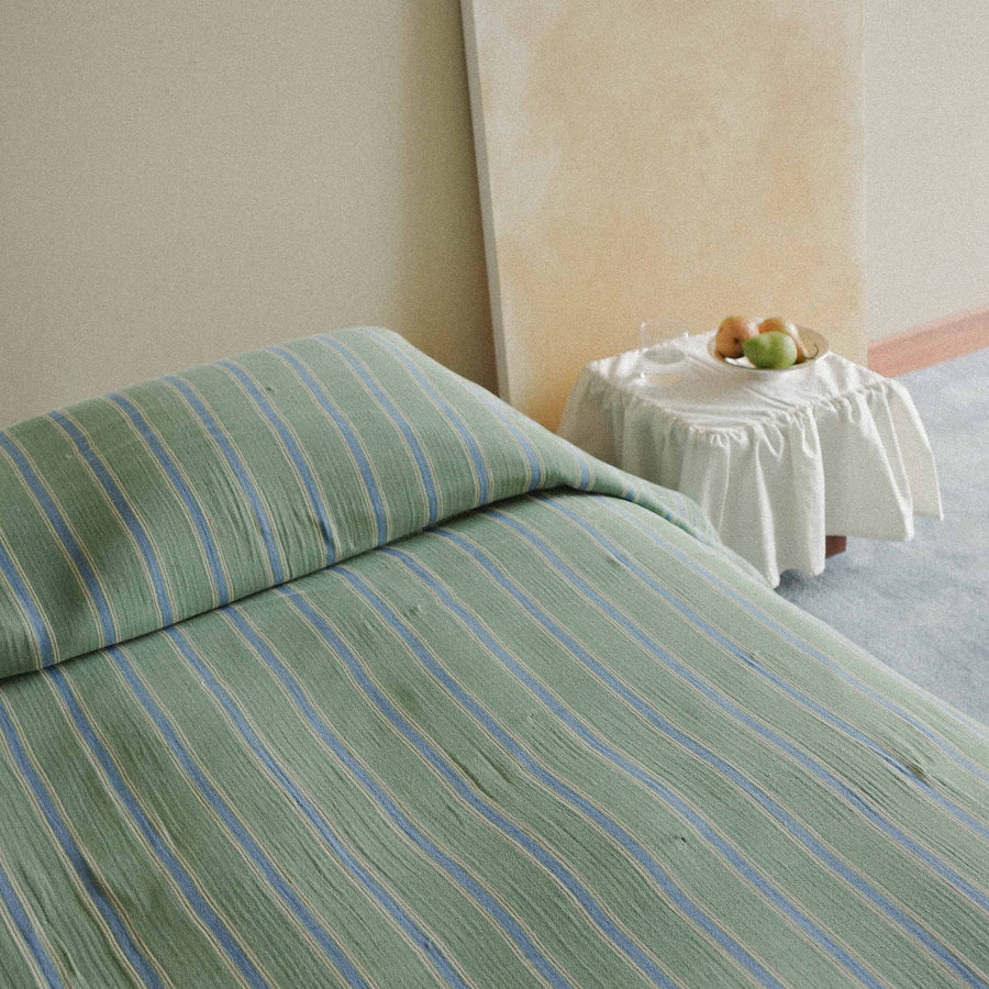 Green and blue striped bedspread on a bed with a side table in the background.