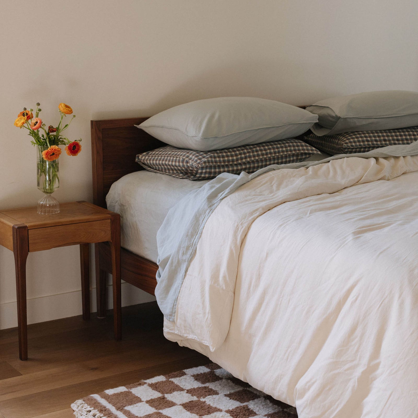 A bed dressed with Mist sheets and an Oat duvet cover. There are Mist pillows stacked neatly atop plaid pillowcases
