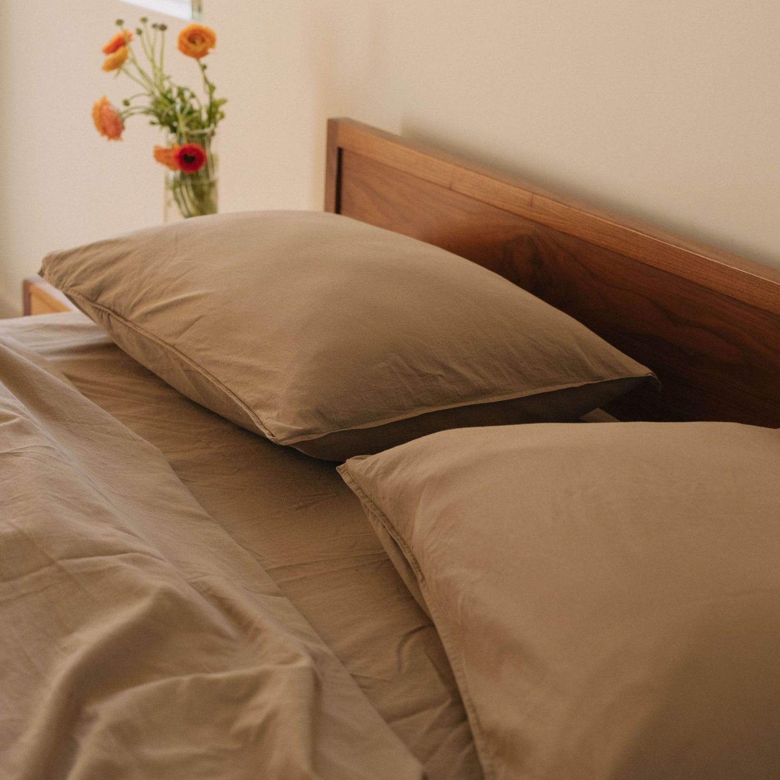 Close up of the head of a bed with beige pillows and sheets against a wood headboard with a vase of flowers in the background