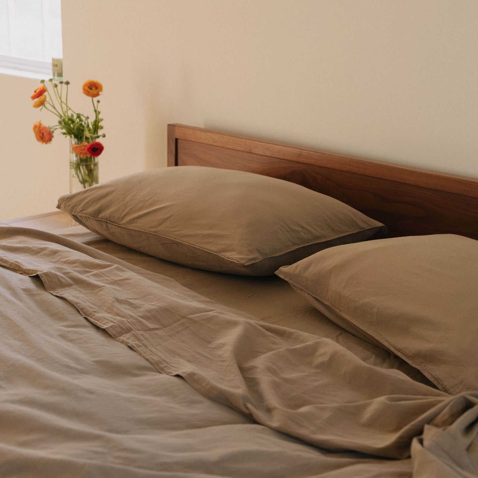 Bed photographed at an angle with Dune / brown sheets and two pillows in a bedroom setting
