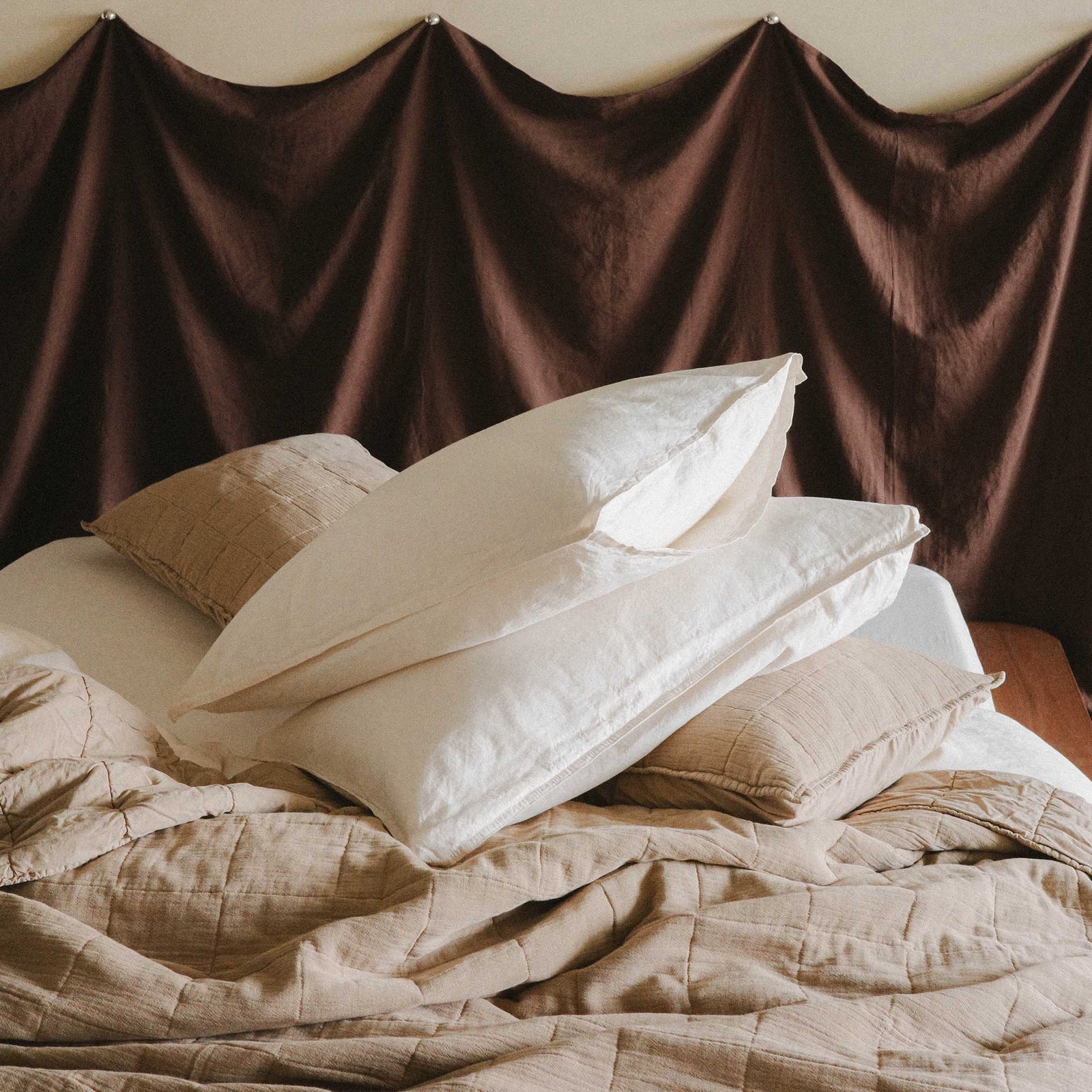 Photo of a bed with a pile of pillows, two white / cream over beige / brown quilted pillowcases laying over a beige / brown quilt 