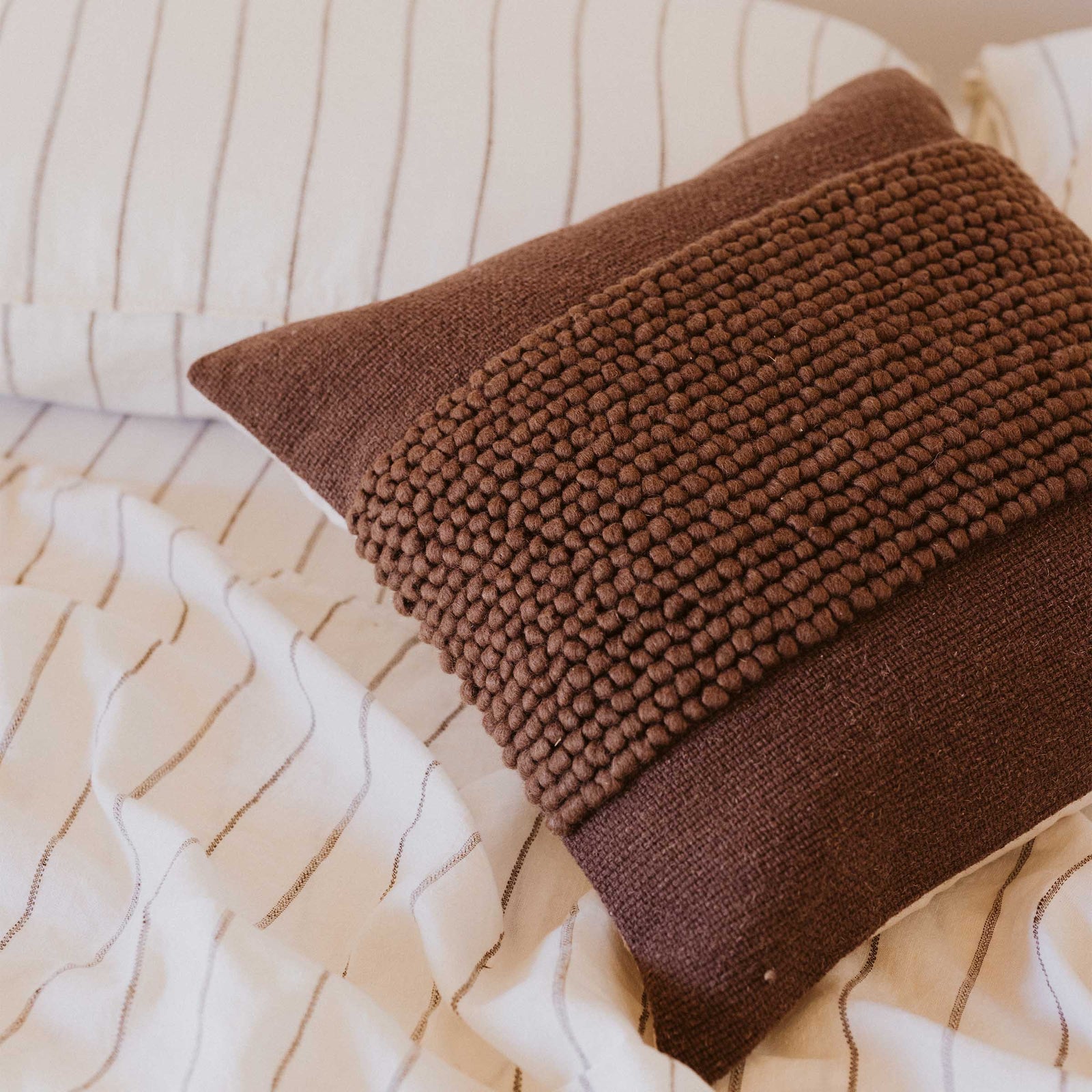 A solid brown throw pillow with a textured center design on a bed with striped sheets.