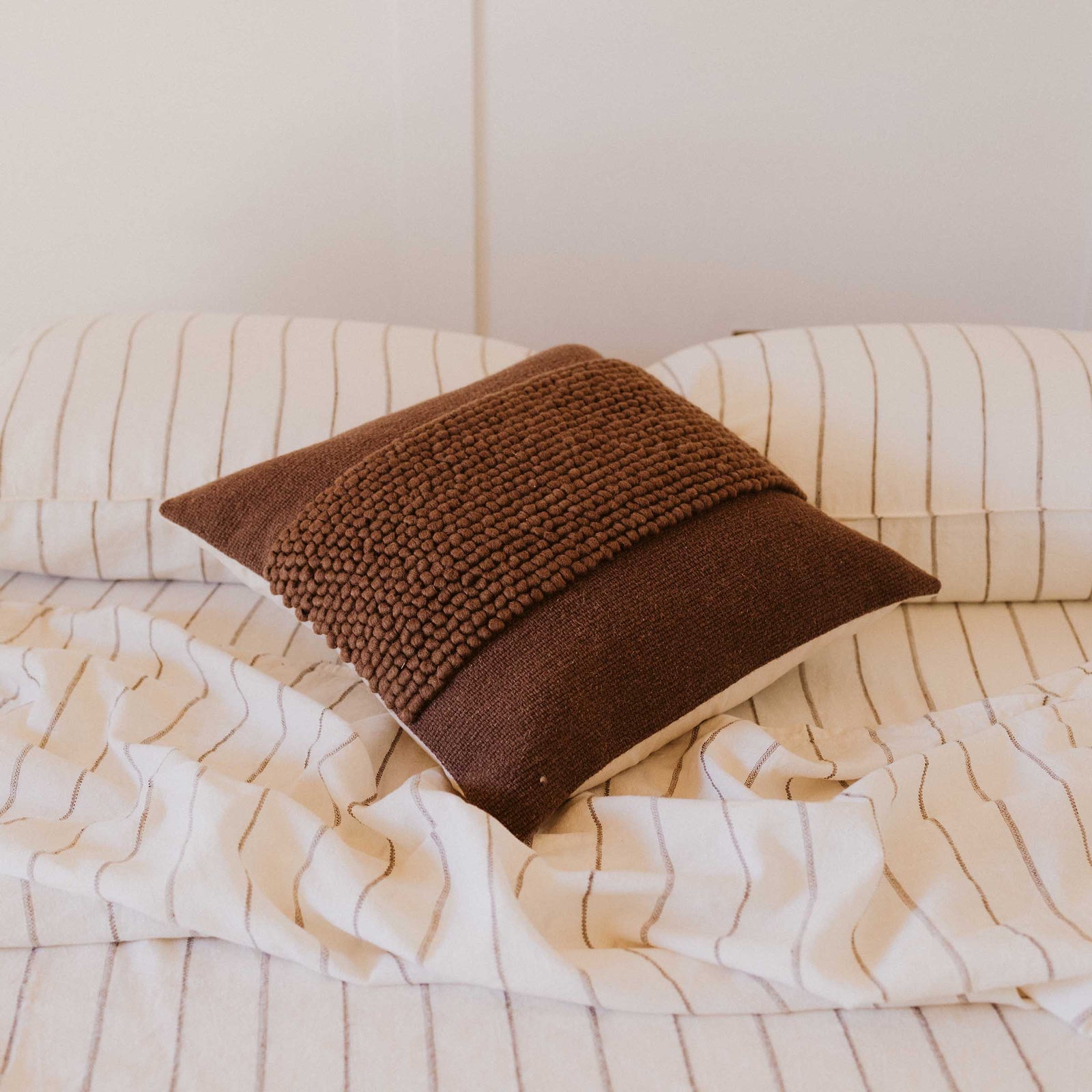 A brown throw pillow with a textured center design and a cream canvas back panel on a bed with striped sheets.