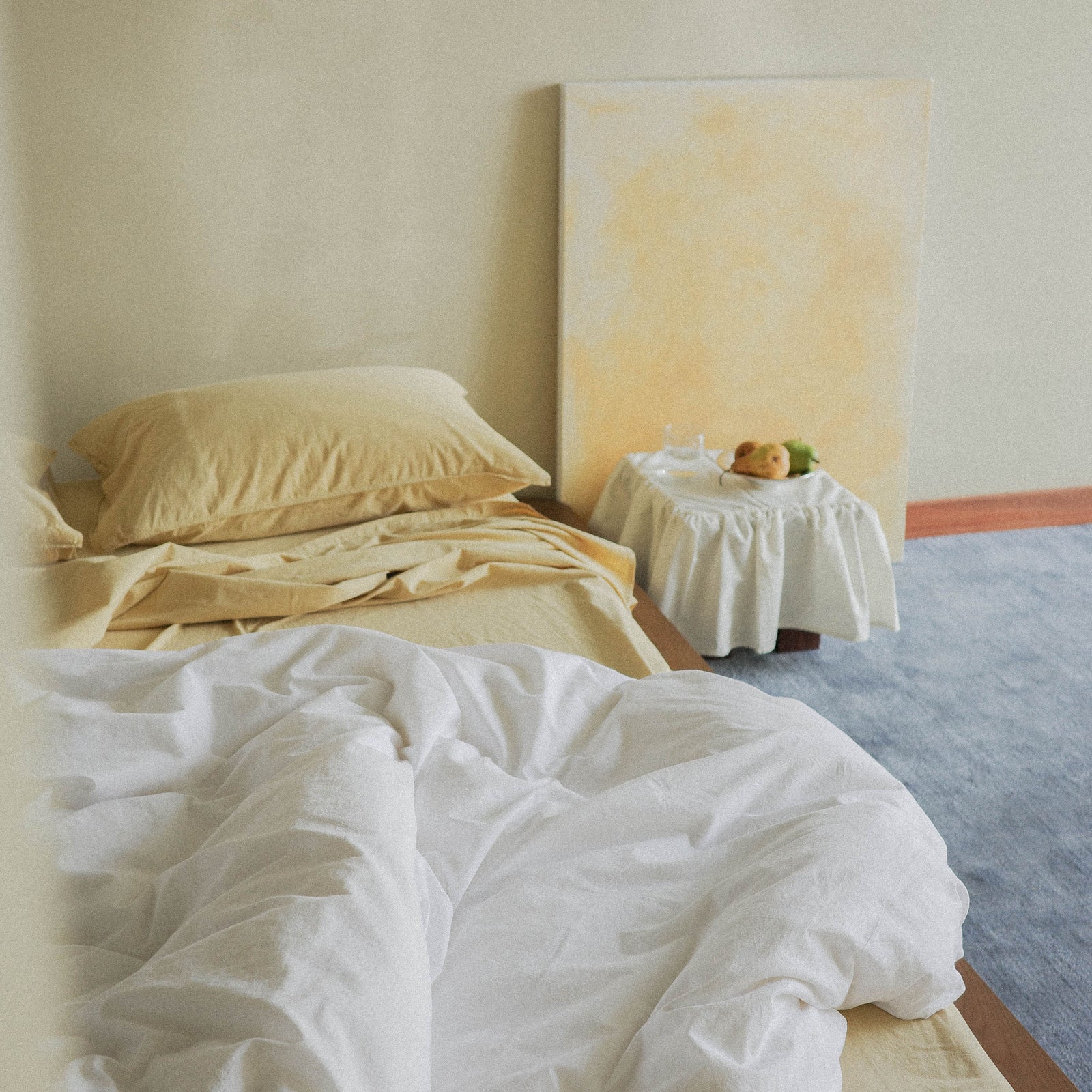 White duvet cover over yellow sheets and pillows on a bed next to a small nightstand with a fruit bowl in the corner.