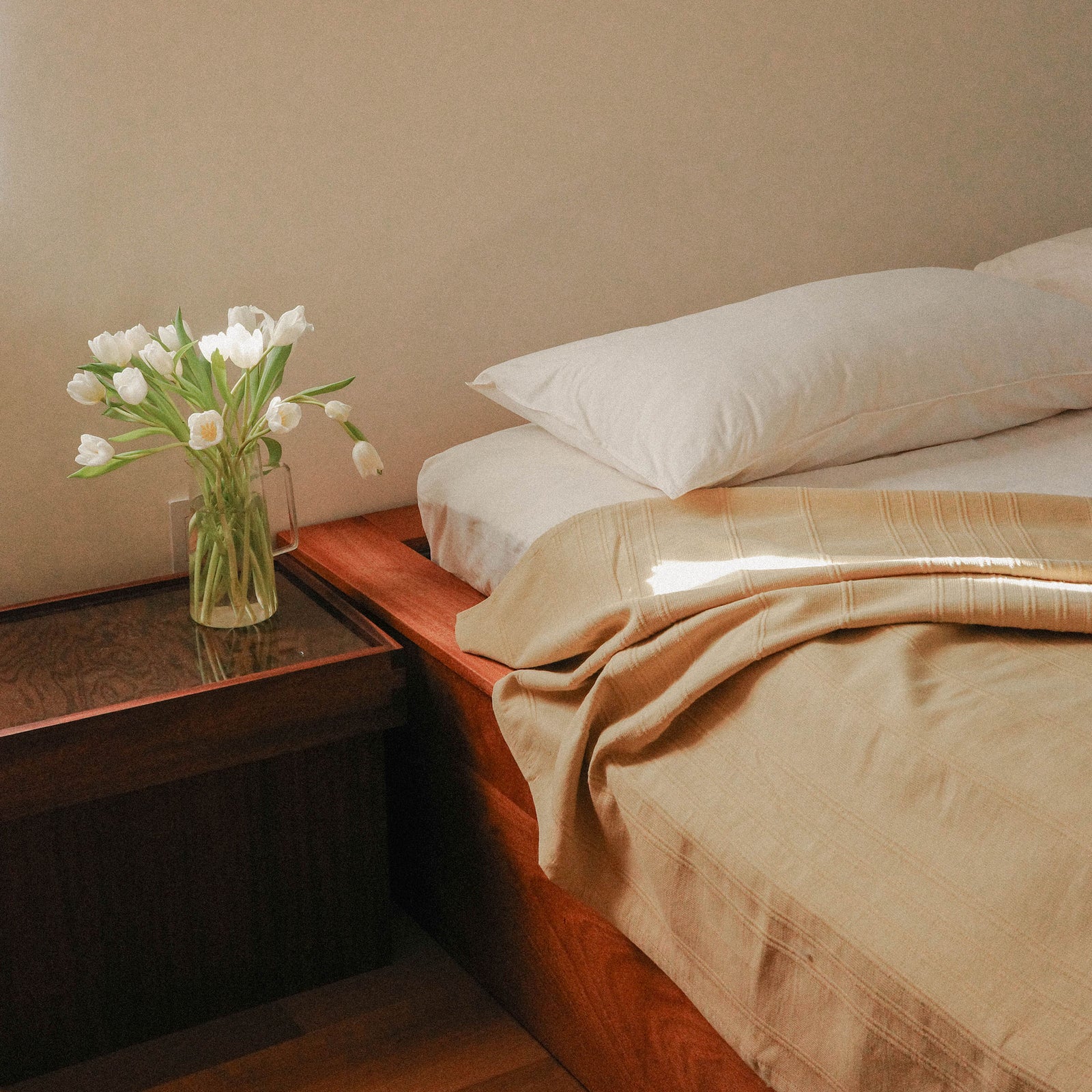 Bedroom with a bed dressed with white and beige sheets and a vase of flowers on a nightstand.