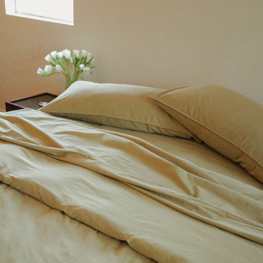 Green sheet set on a bed with a side table and flowers in the background.