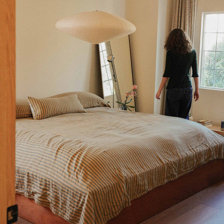 Person standing by a bed with a coverlet on it, in a room with a lamp and window and mirror.