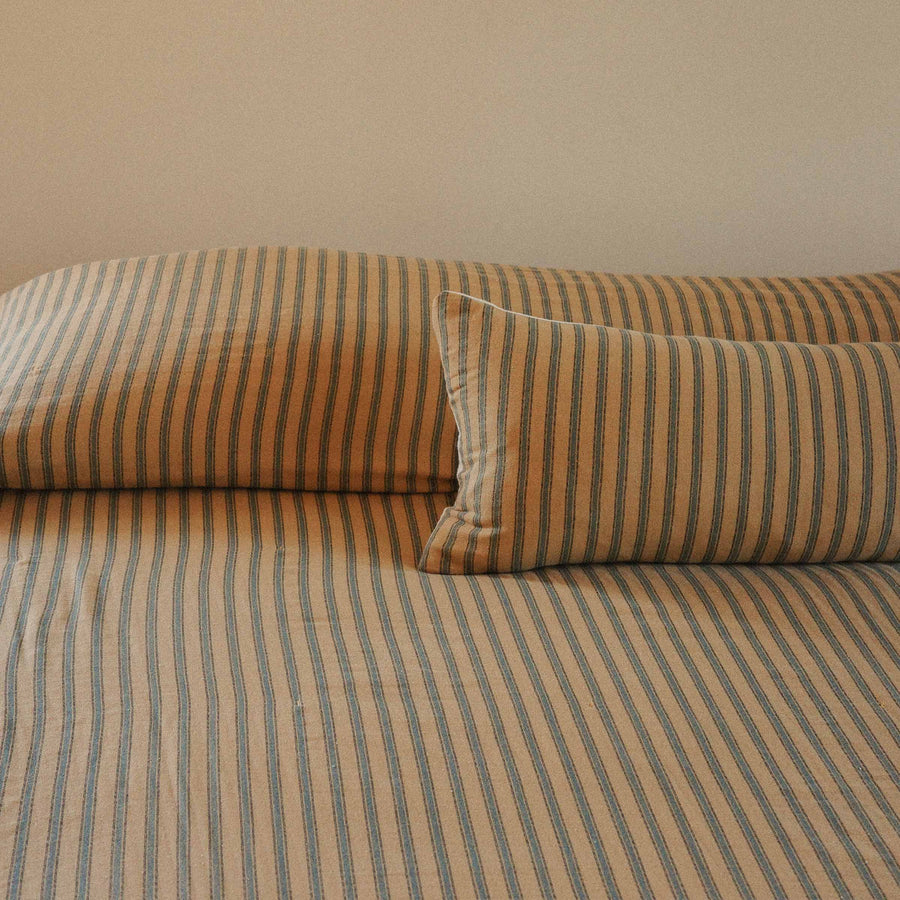 Striped coverlet with folded over two pillows with a lumbar pillow in front, all in the same pattern in front of a beige background