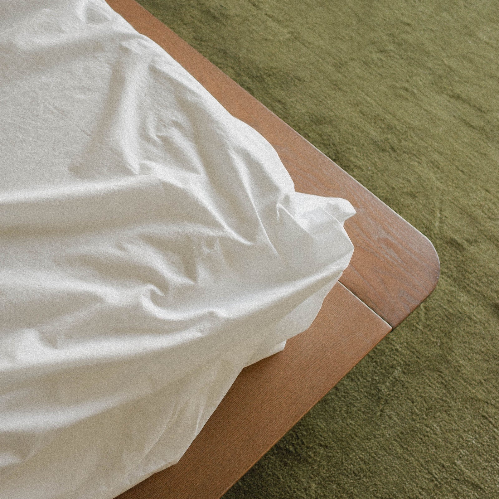 White flat sheet draped over a wooden bedframe on a green carpeted floor
