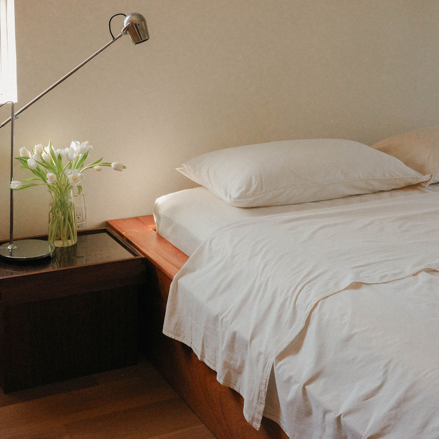 Cream sheets with pillows on a bed next to a wooden nightstand with vase of white flowers.