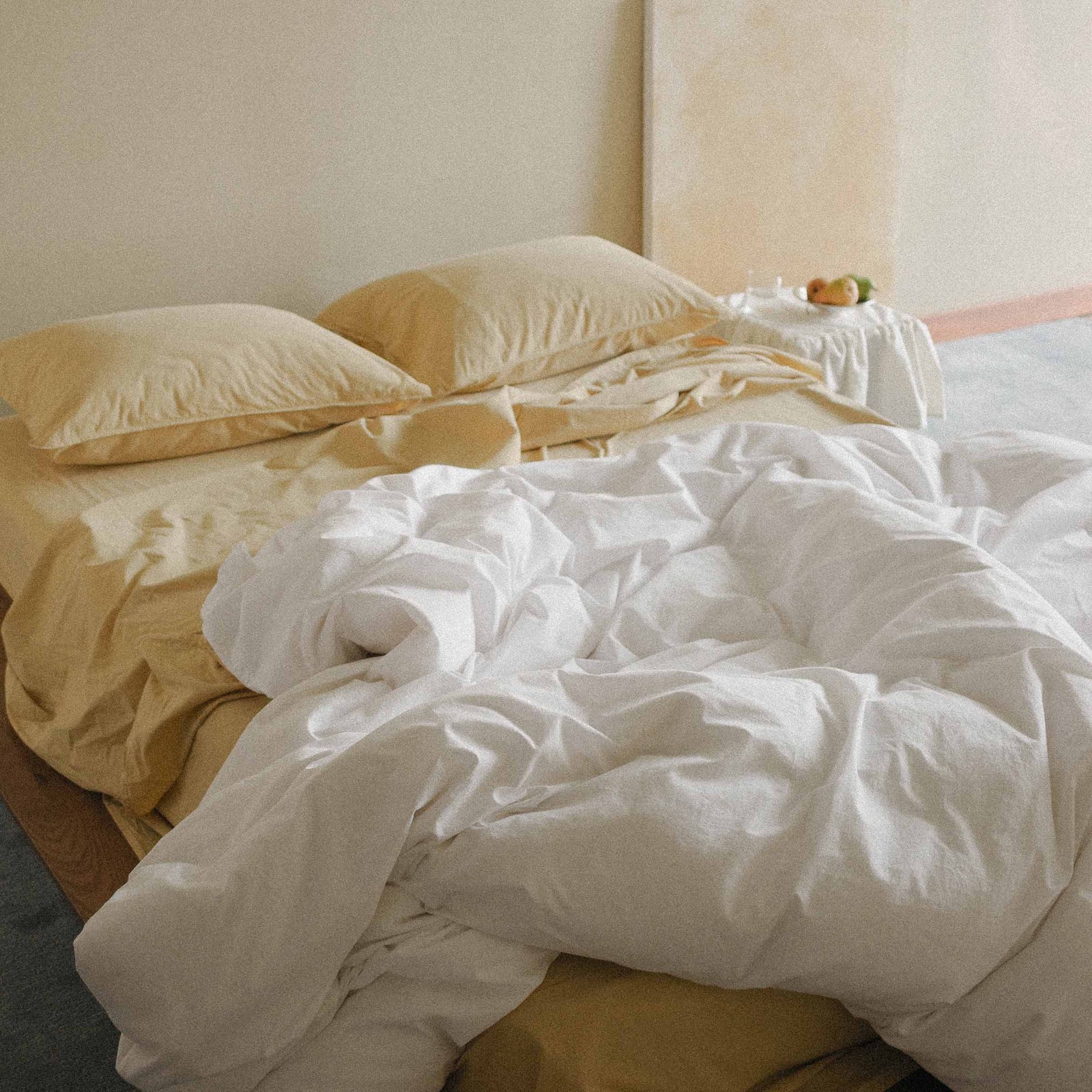 Unmade bed with white duvet cover and yellow sheets and pillows against a neutral wall.