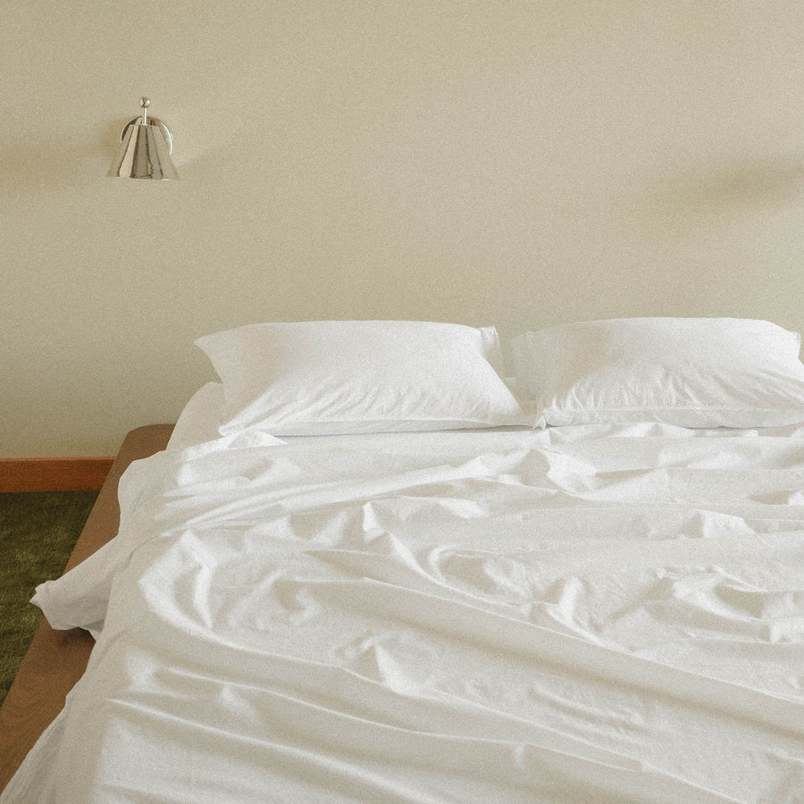 Bed with white bedding and pillows against a beige wall with a silver sconce and green carpet.