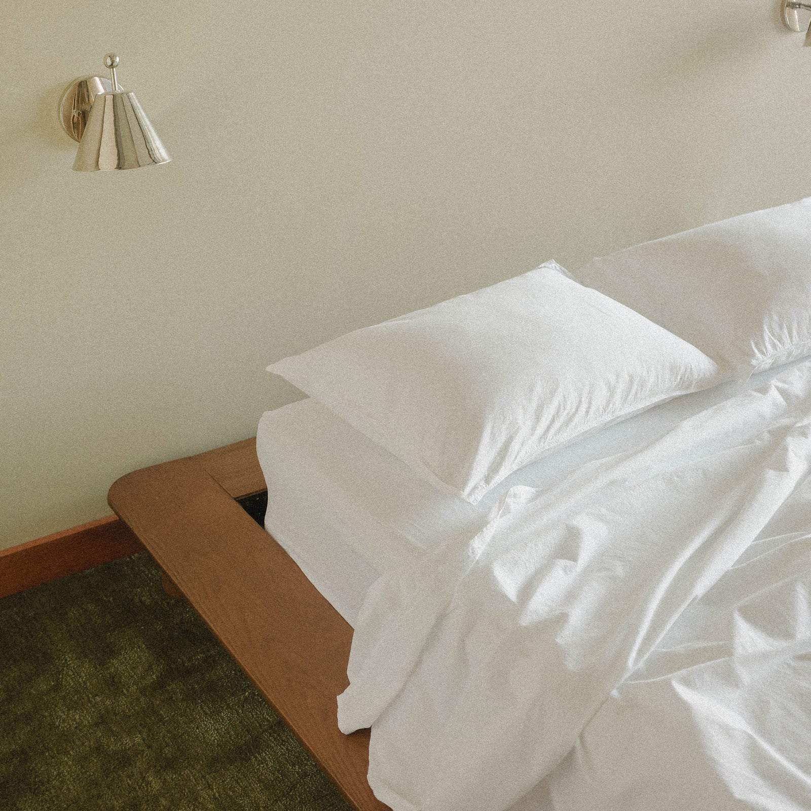 Bed with white sheets in a room with a silver sconce on the wall and green carpet.