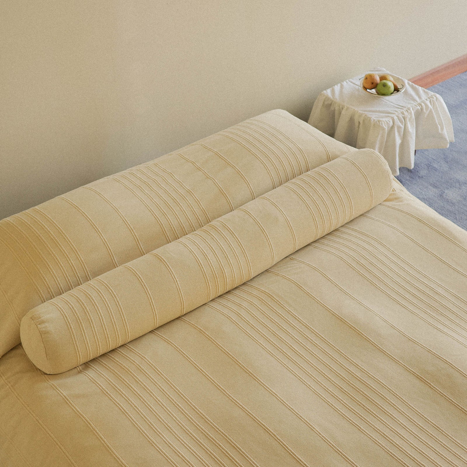 Morrow Soft Goods Devi Coverlet and Devi Bolster in Maiz - Neatly made bed with a beige / yellow coverlet and matching bolster in a bedroom setting beside a small table with a bowl of fruit